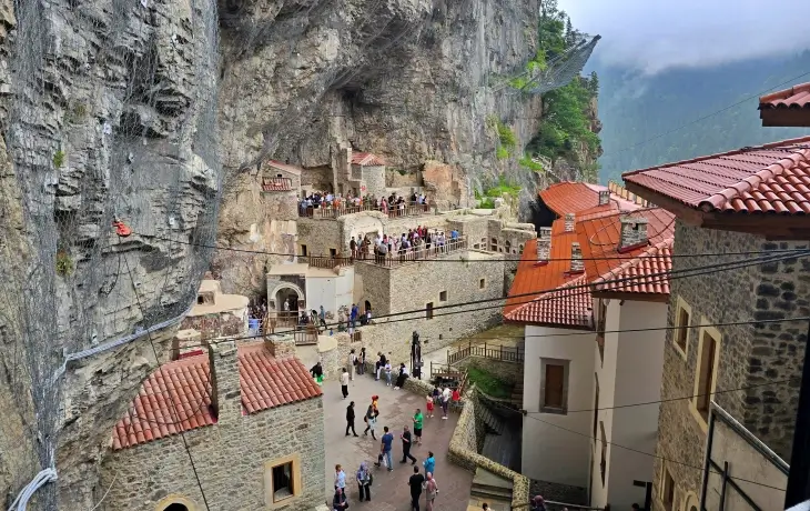 Trabzon Sümela Manastırı avlu ve teras bölümü; ana kaya kütlesine oyulmuş yapılar, kırmızı kiremitli keşiş hücreleri ve ziyaretçi kalabalığıyla birlikte kompleksin genel görünümü.
